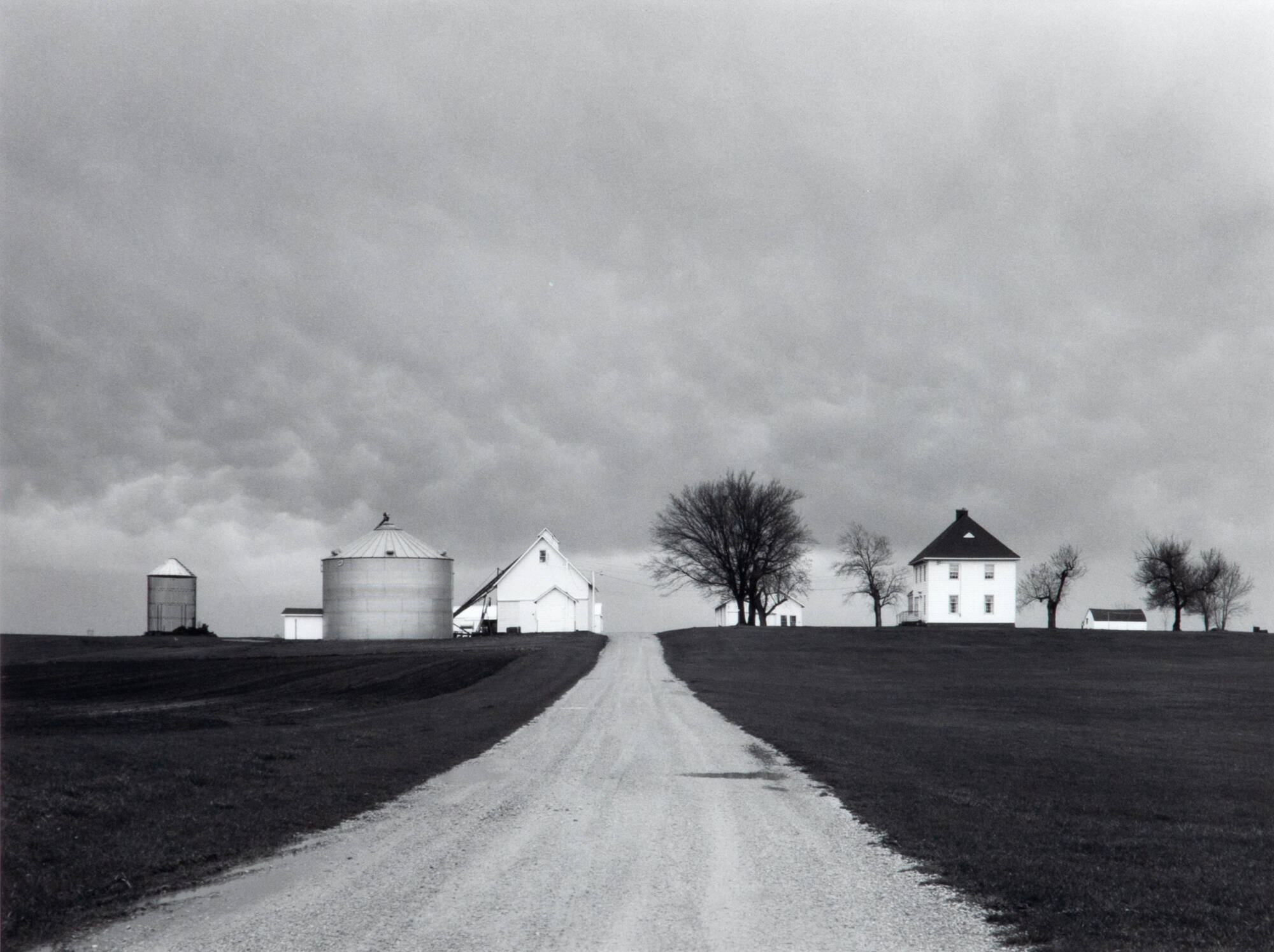 Black and white photograph of a farm landscape scene with a dirt road running straight up center; grain mill and white house; sky is dark, storm brewing.